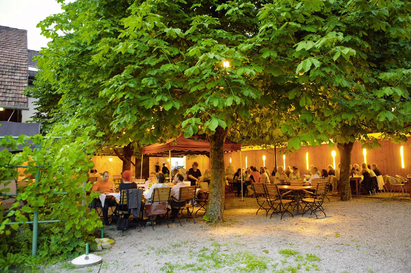 Der Gastgarten am Abend: besetzte Tische unter Kastanienbäumen, Fackeln an der Holzwand