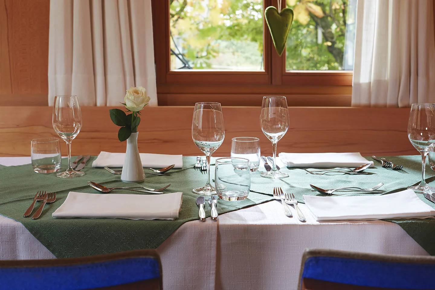 Gedeckter Tisch am Fenster mit weißer Rose, grünem Läufer und einem grünen Stoffherz am Fensterrahmen